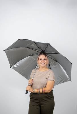 Lächelnde Frau mit tätowiertem Unterarm hält einen geöffneten silbergrauen Regenschirm vor neutralem Hintergrund.