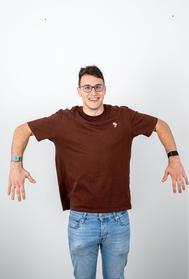 Junger Mann mit Brille und braunem T-Shirt hebt die Arme seitlich an, lächelt in die Kamera und steht vor neutralem, hellem Hintergrund.