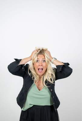 Blonde Frau mit weit aufgerissenen Augen fasst sich mit beiden Händen an den Kopf und zeigt einen Gesichtsausdruck der Überraschung oder Überforderung.