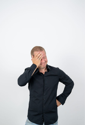 Mann mittleren Alters in schwarzem Hemd steht vor neutralem Hintergrund und hält sich mit einem leicht genervten oder beschämten Gesichtsausdruck die Hand an die Stirn.