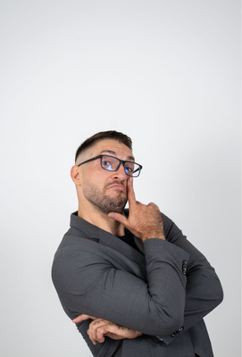 Mann mit Brille und kurzem Bart in grauem Sakko legt nachdenklich einen Finger an die Wange und blickt zur Seite.