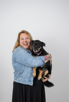 Lächelnde Frau mit blondem Haar und Jeansjacke hält einen kleinen schwarzen Hund mit braunen Abzeichen und rosa Halsband im Arm, sichtbares Blumen-Tattoo am rechten Arm.