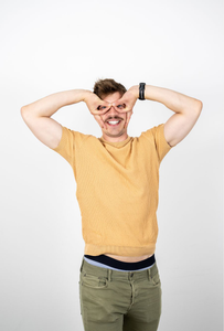 Mann mit Schnurrbart in gelbem T-Shirt macht mit den Händen eine spielerische Geste vor dem Gesicht, die eine Brille imitiert, und lächelt breit in die Kamera.