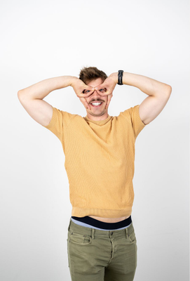 Mann mit Schnurrbart in gelbem T-Shirt macht mit den Händen eine spielerische Geste vor dem Gesicht, die eine Brille imitiert, und lächelt breit in die Kamera.