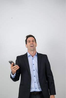 Mann im Business-Outfit mit Smartphone in der rechten Hand und kabellosen Kopfhörern, lächelnd und in kommunikativer Haltung vor neutralem Hintergrund.