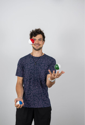 Lächelnder junger Mann mit lockigen Haaren jongliert drei bunte Bälle vor neutralem Hintergrund.