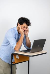 Mann mittleren Alters steht an einem kleinen Schreibtisch und stützt den Kopf in die Hände, während er mit angespanntem Gesichtsausdruck auf einen Laptop blickt.