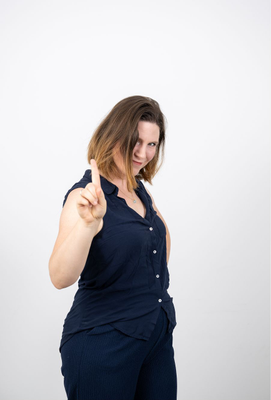 Frau mit schulterlangem Haar und dunkelblauer Kleidung hebt den Zeigefinger mahnend, während sie leicht lächelt und direkt in die Kamera blickt.