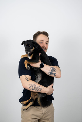 Mann mit Tätowierungen auf beiden Armen hält einen kleinen schwarz-braunen Hund und schaut lächelnd in die Kamera vor neutralem Hintergrund.