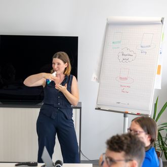 Frau hält Präsentation an einem Flipchart, während eine Gruppe von Teilnehmenden aktiv zuhört und Notizen an Laptops macht.