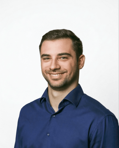 Ein lächelnder Mann in einem blauen Hemd vor einem neutralen Hintergrund.