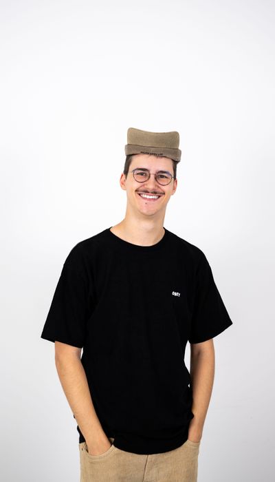 Person mit schwarzem T-Shirt und beige Hut, lächelnd mit Händen in den Hosentaschen, vor weißem Hintergrund.