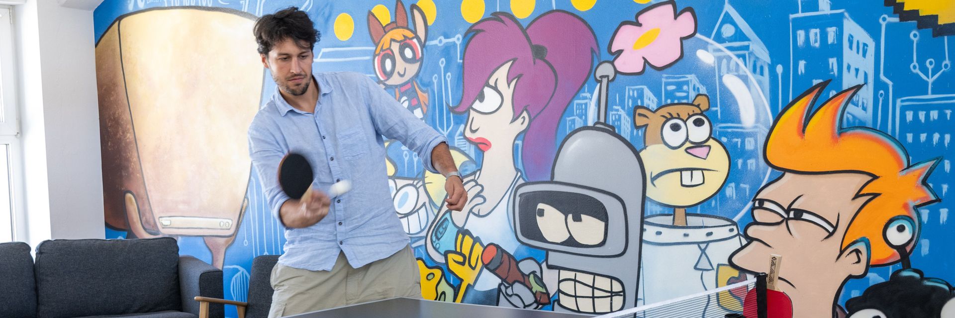 Person spielt Tischtennis in einem Büro, das eine bunte Wand mit Graffiti-Kunst zeigt, darunter bekannte animierte Figuren.
