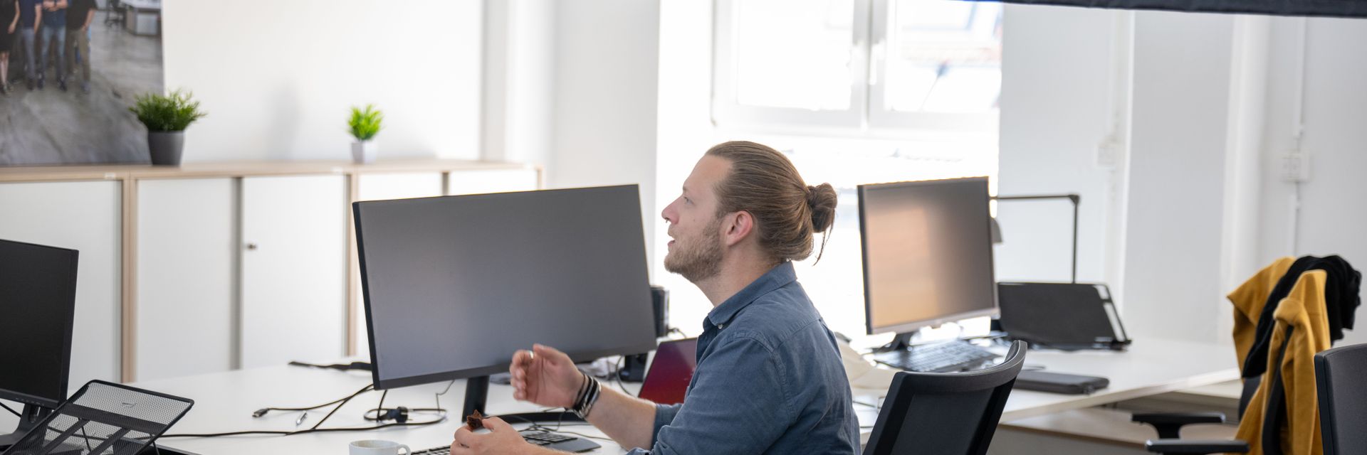 Person sitzt an einem Schreibtisch in einem modernen Büro, arbeitet am Computer und hält Gebäck in der Hand. Im Hintergrund sind weitere Arbeitsplätze und Pflanzen zu sehen.