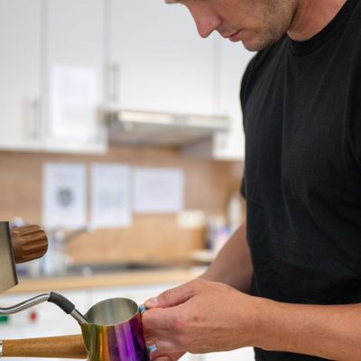 Mann in schwarzem T-Shirt schäumt Milch in einem bunten Kännchen mit einer Espressomaschine auf.