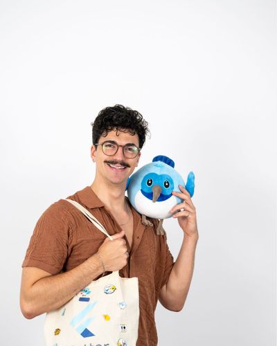 Person mit lockigem Haar und Schnurrbart, die eine Stofftasche mit dem Flutter-Logo trägt und ein blaues Stofftier lächelnd an der rechten Schulter hält.