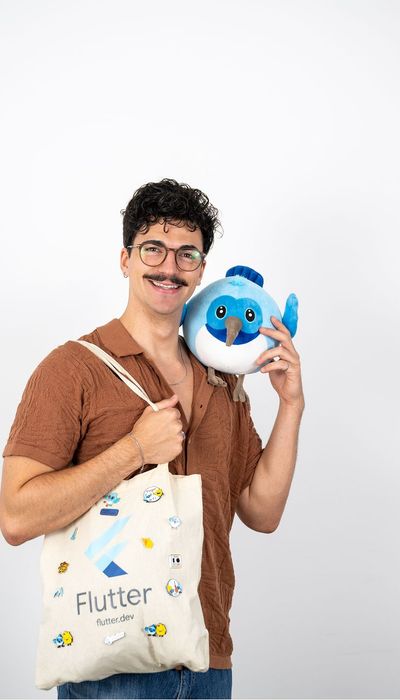 Person mit lockigem Haar und Schnurrbart, die eine Stofftasche mit dem Flutter-Logo trägt und ein blaues Stofftier lächelnd an der rechten Schulter hält.