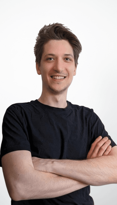 Person in schwarzem T-Shirt lächelt mit verschränkten Armen vor einem neutralen Hintergrund.