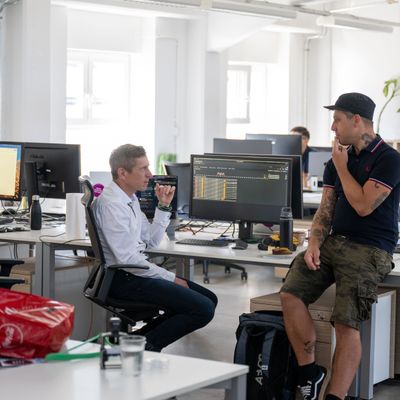 Ein Mann im weißen Hemd sitzt am Schreibtisch mit Monitoren, während ein anderer Mann in Freizeitkleidung auf dem Schreibtisch sitzt, beide im Gespräch.