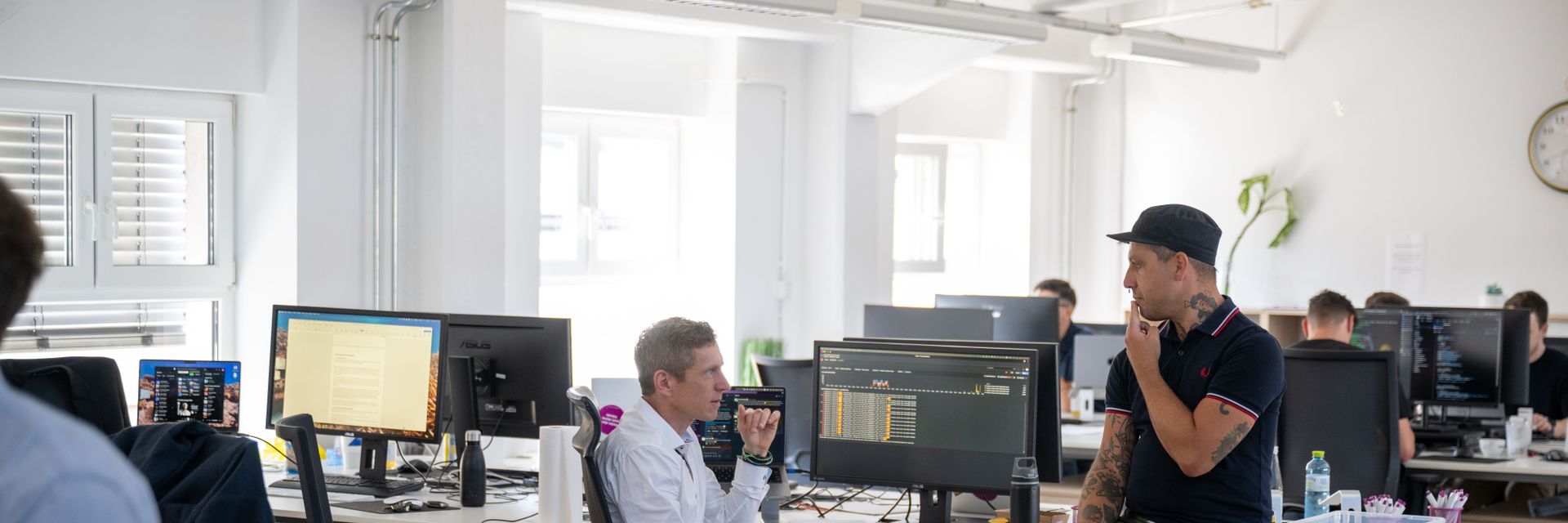 Ein Mann im weißen Hemd sitzt am Schreibtisch mit Monitoren, während ein anderer Mann in Freizeitkleidung auf dem Schreibtisch sitzt, beide im Gespräch.