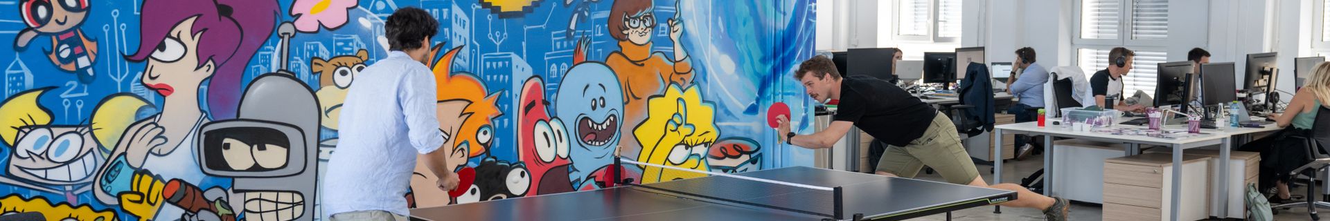 Zwei Personen spielen Tischtennis in einem Büro mit buntem Wandgemälde von Cartoon-Figuren. Im Hintergrund arbeiten Mitarbeiter an Schreibtischen.