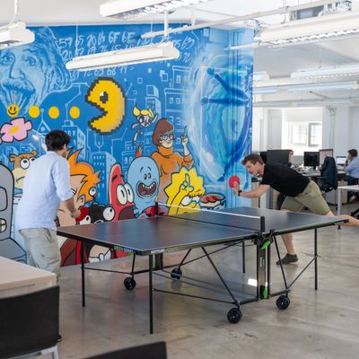 Zwei Personen spielen Tischtennis in einem Büro mit buntem Wandgemälde von Cartoon-Figuren. Im Hintergrund arbeiten Mitarbeiter an Schreibtischen.
