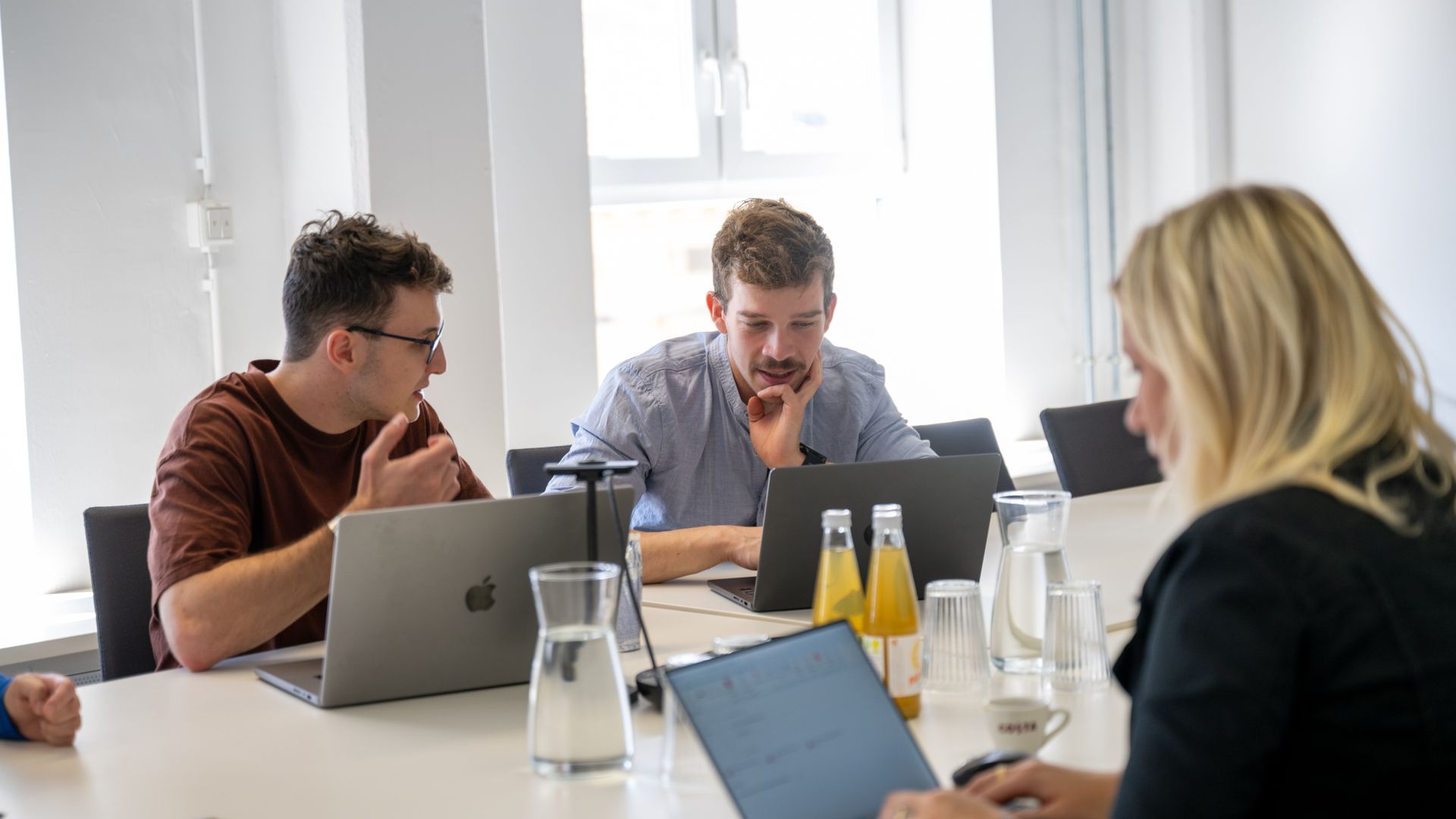 Drei Personen in einem modernen Besprechungsraum arbeiten gemeinsam an Laptops, während sie sich angeregt unterhalten. Auf dem Tisch stehen Getränke und Wasserkrüge.