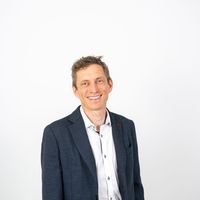 Junger Mann in einem dunklen Blazer und weißem Hemd, lächelnd vor einem hellen Hintergrund stehend.