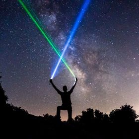 Person hält zwei Laser unter sternklarem Himmel, mit der Milchstraße im Hintergrund.