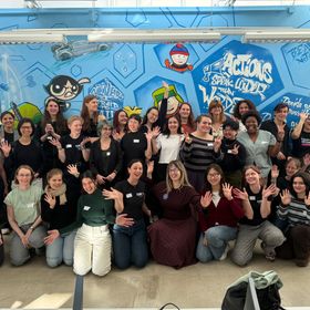 Gruppenfoto von 38 Frauen, die sich vor einer bunten Wand mit Graffiti versammeln und lächeln. Viele Teilnehmerinnen haben die Hände gehoben, um zu winken oder Zeichen zu machen. Die Wand zeigt verschiedene Charaktere und inspirierende Sprüche, die die Atmosphäre von Gemeinschaft und Unterstützung vermitteln.