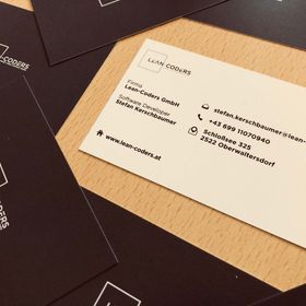 Visitenkarte von Lean-Coders mit Kontaktinformationen auf einem Tisch, umgeben von schwarzen Visitenkarten mit Unternehmenslogo.