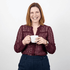 Frau mit schulterlangen, braunen Haaren lächelt und hält eine weiße Tasse in der Hand. Sie trägt eine bordeauxfarbene Bluse aus Spitze und eine dunkelblaue Hose. Der Hintergrund ist neutral und hell.