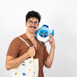 Person mit lockigem Haar und Schnurrbart, die eine Stofftasche mit dem Flutter-Logo trägt und ein blaues Stofftier lächelnd an der rechten Schulter hält.