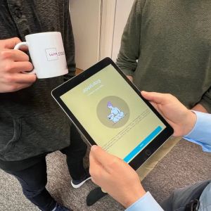 Person hält ein Tablet mit einer geöffneten App, während zwei andere Personen daneben stehen; eine hält eine Kaffeetasse.