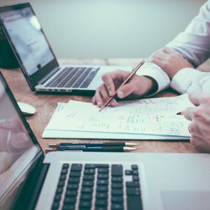 Zwei Personen arbeiten an einem Schreibtisch mit Laptops und Papieren, wobei eine Person mit einem Stift auf ein Dokument zeigt.