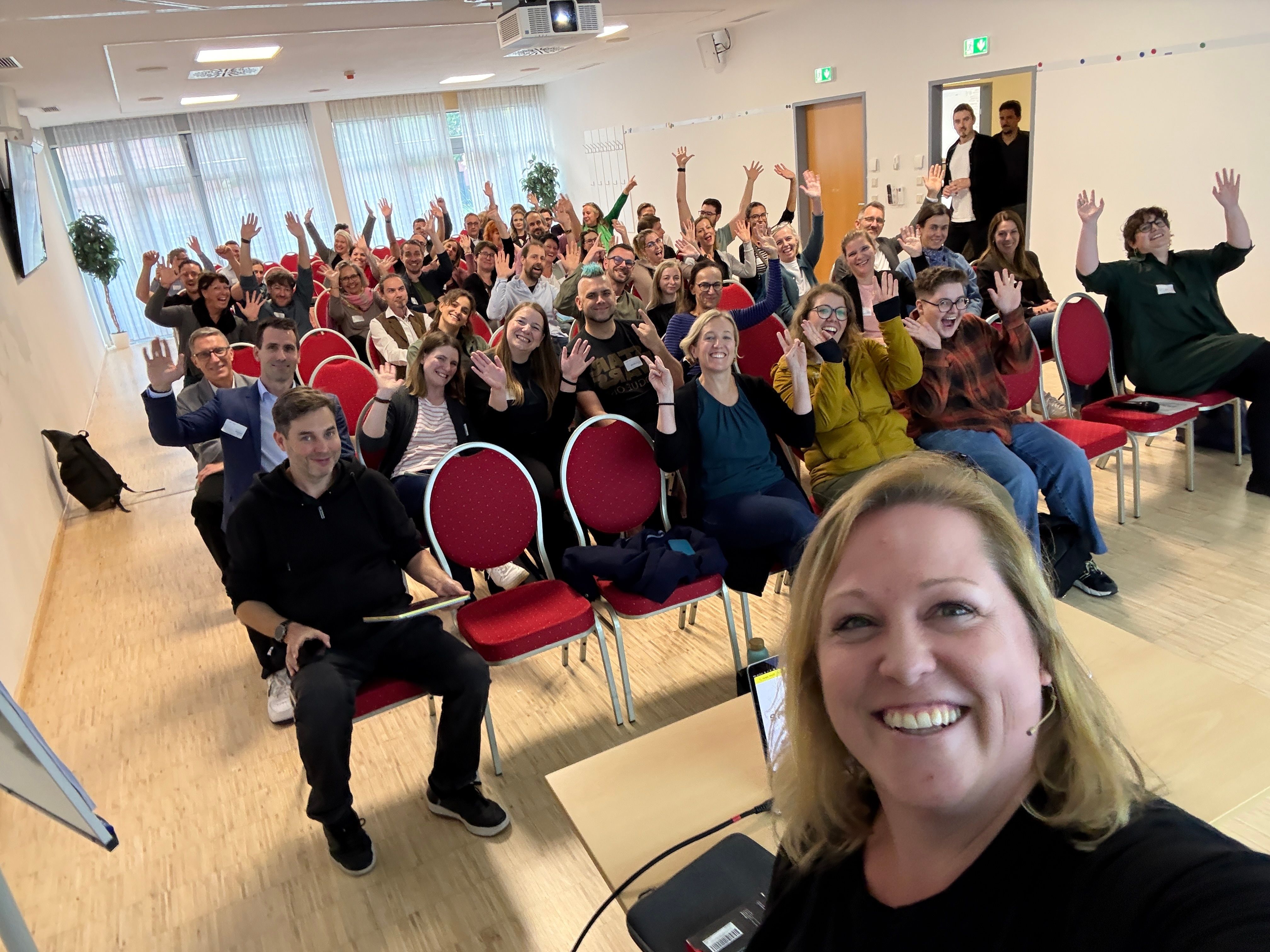 Gruppenfoto von Teilnehmern einer Veranstaltung, die in einem Seminarraum sitzen und der Kamera zuwinken. Im Vordergrund ist eine lächelnde Frau zu sehen, die das Foto aufnimmt. Die Mehrheit der Anwesenden trägt lässige Kleidung und zeigt eine positive, freundliche Atmosphäre. Rote Stühle und ein Tisch mit technischer Ausstattung sind im Bild sichtbar.