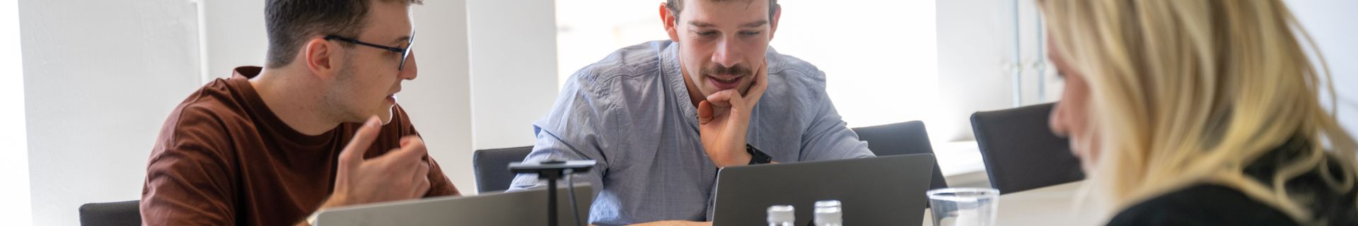 Drei Personen in einem modernen Besprechungsraum arbeiten gemeinsam an Laptops, während sie sich angeregt unterhalten. Auf dem Tisch stehen Getränke und Wasserkrüge.