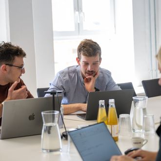 Personen bei einem Meeting diskutieren an einem Konferenztisch, auf dem Laptops und Getränkeflaschen stehen.