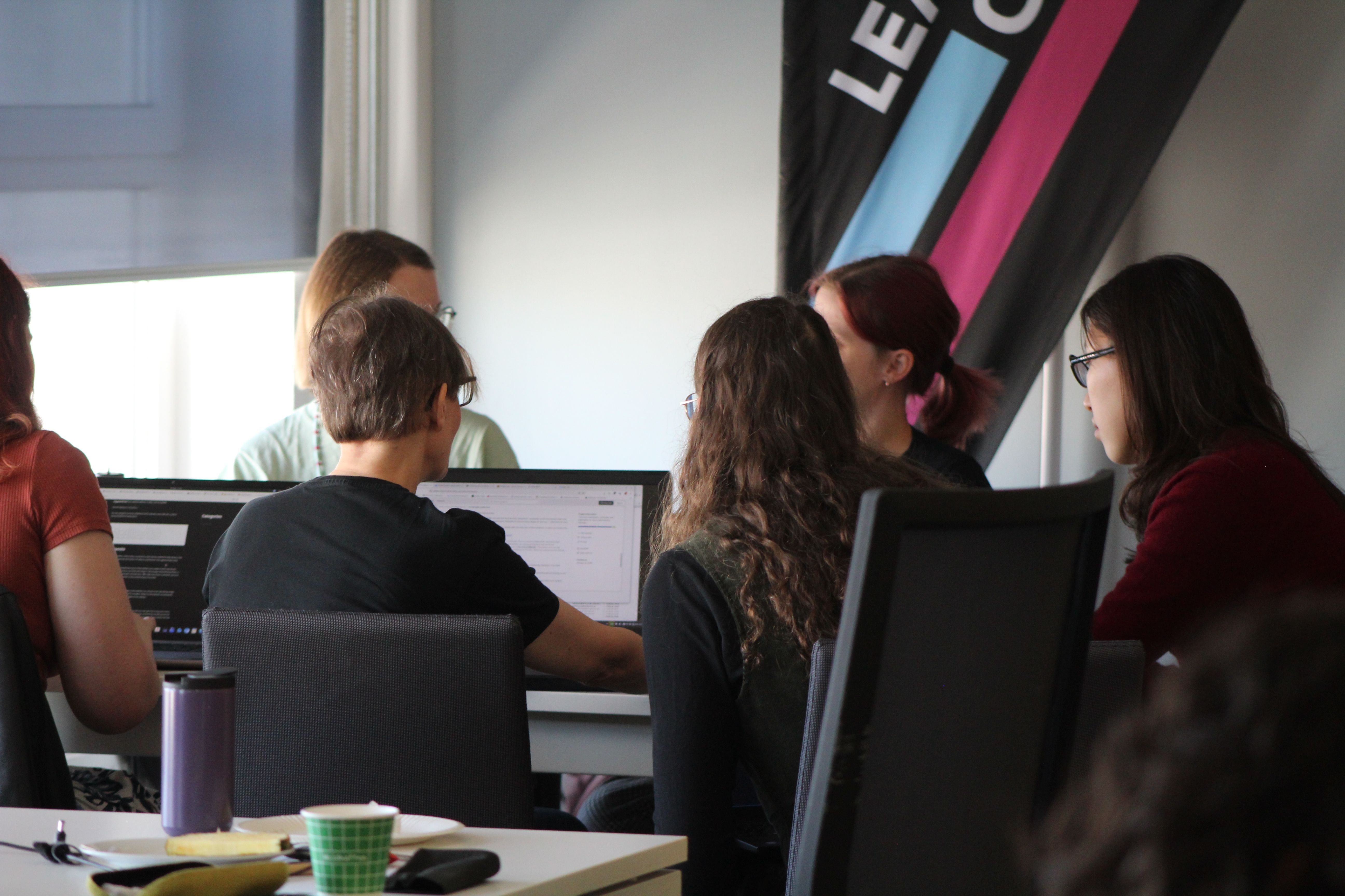 Gesprächsrunde in einem modernen Büro, bei der mehrere Personen an Computern arbeiten. Im Vordergrund sind zwei Personen zu sehen, die konzentriert auf ihre Bildschirme blicken, während im Hintergrund weitere Teilnehmer an einer Diskussion teilnehmen. Auf dem Tisch befinden sich Tassen und Snacks.