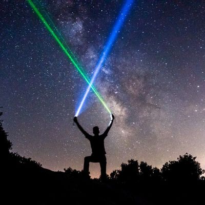 Person hält zwei Laser unter sternklarem Himmel, mit der Milchstraße im Hintergrund.