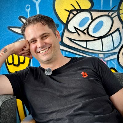 Lächelnder Mann mit kurzer Haarfrisur, der entspannt auf einem Sessel sitzt und seinen Kopf auf die Hand stützt. Im Hintergrund ist ein bunter Graffiti mit einem cartoonhaften Charakter zu sehen.