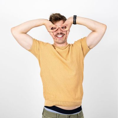 Mann in einem hellbraunen T-Shirt, der lächelnd eine Brille mit den Händen andeutet.