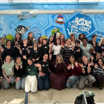 Gruppenfoto von 38 Frauen, die sich vor einer bunten Wand mit Graffiti versammeln und lächeln. Viele Teilnehmerinnen haben die Hände gehoben, um zu winken oder Zeichen zu machen. Die Wand zeigt verschiedene Charaktere und inspirierende Sprüche, die die Atmosphäre von Gemeinschaft und Unterstützung vermitteln.