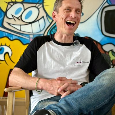 Lachender Mann mit kurzen, grauen Haaren, der in einem gemütlichen Sessel sitzt. Er trägt ein weißes T-Shirt mit schwarzem Ärmel und Jeans. Im Hintergrund ist eine bunte Wandmalerei mit cartoonhaften Figuren zu sehen.