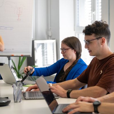 Kollegen in einem Meetingraum, die an Laptops arbeiten, während eine Frau am Whiteboard präsentiert.