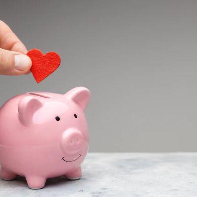 Hand legt ein rotes Herz in ein rosa Sparschwein.