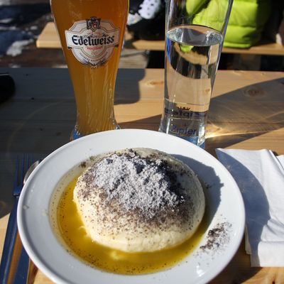 Germknödel mit Mohnzucker und flüssiger Butter auf einem weißen Teller, daneben ein Glas Weizenbier und ein Glas Wasser auf einem Holztisch im Freien.
