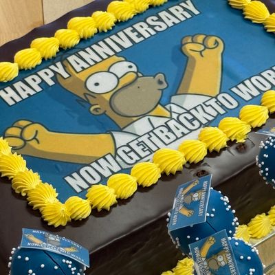 Rechteckige Schokoladentorte mit gelber Verzierung und Aufschrift „Happy Anniversary, Now Get Back to Work“ auf einer bedruckten Zuckerfolie mit einer bekannten Cartoon-Figur.