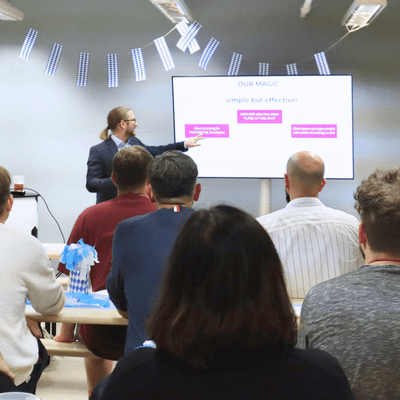 Person hält eine Präsentation vor einer Gruppe von Menschen, die auf Bänken sitzen, während auf einem Bildschirm eine Folie mit Text angezeigt wird.