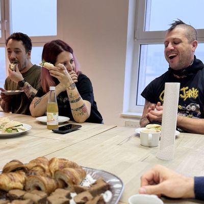 Personen sitzen an einem Tisch und essen Snacks, darunter Gebäck und Sandwiches. Auf dem Tisch steht eine Flasche Mate-Tee.